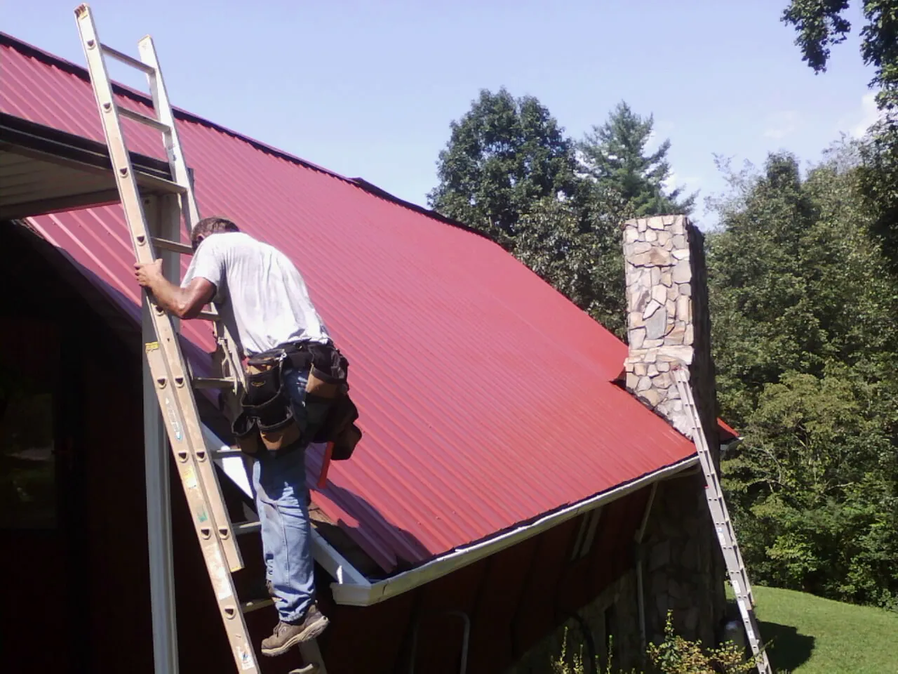 Roofer on a ladder beside a red metal roof and stone chimney — Soffit & Fascia Repair by Roofing Brothers in Medicine Lodge
